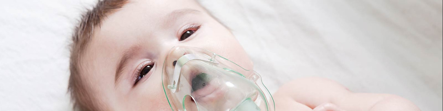 Baby laying in bed with a nebulizer mask delivering liquid medication into a medicated mist.
